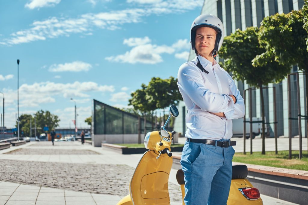 Confident handsome man standing next to a yellow classic italian scooter against a skyscraper.