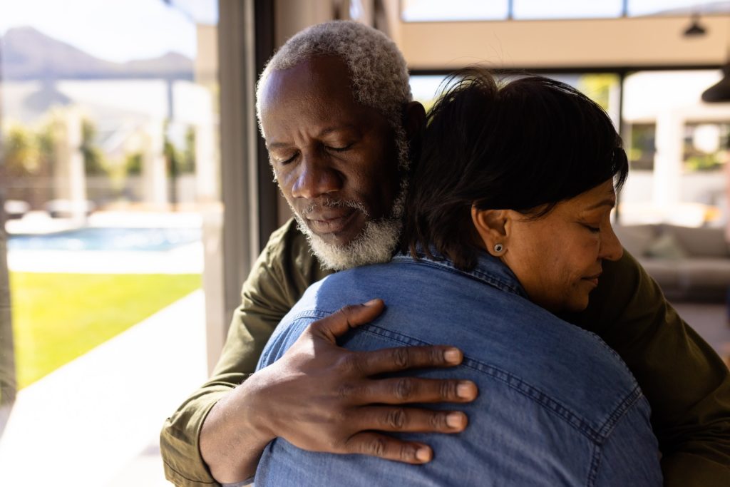 Multiracial senior man with eyes closed embracing woman while standing in nursing home